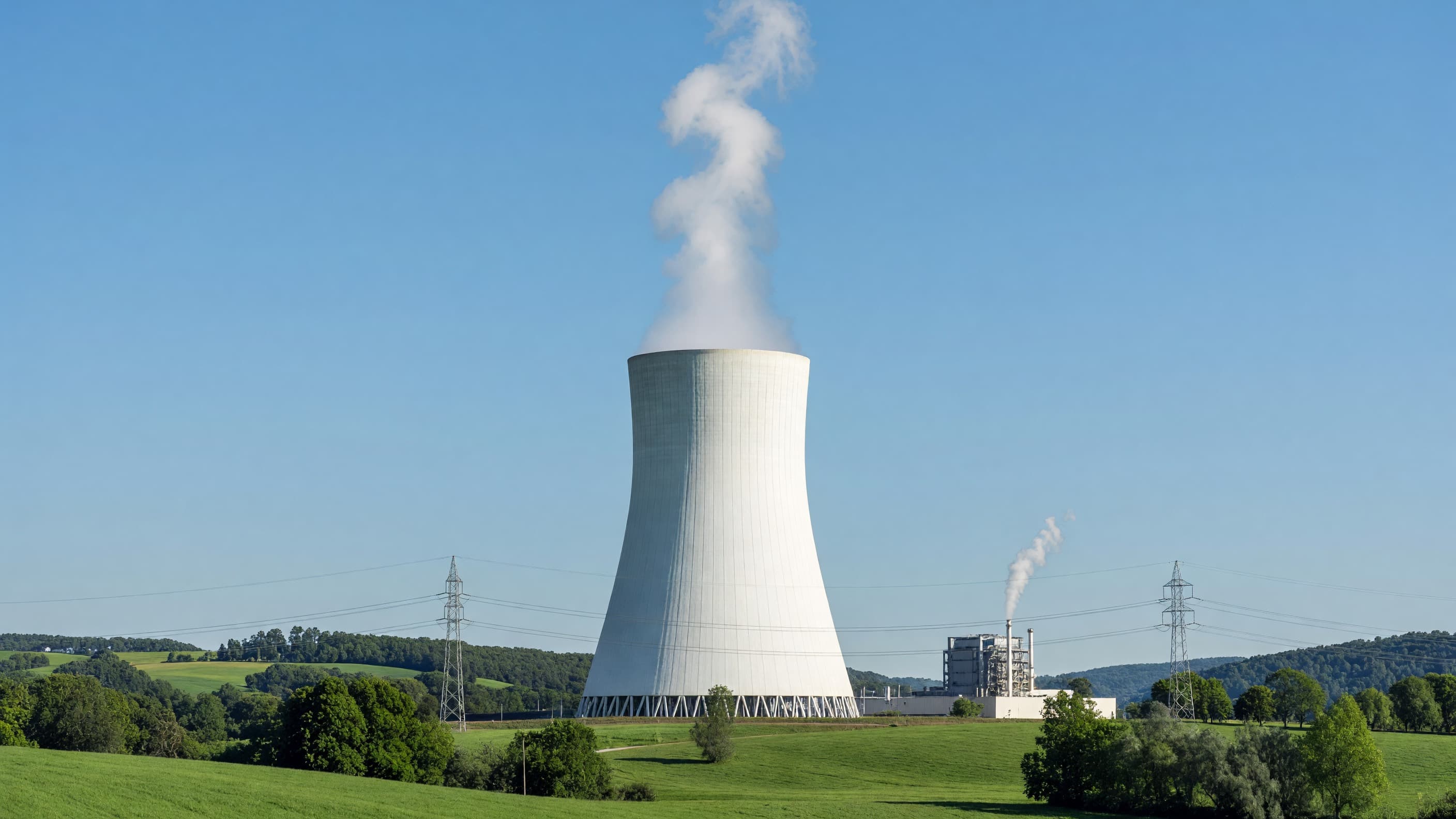A nuclear power plant cooling tower in green farmland under a clear blue sky, with a smaller gas plant beyond, illustrating nuclear versus natural-gas baseload power. Illustration: NNN