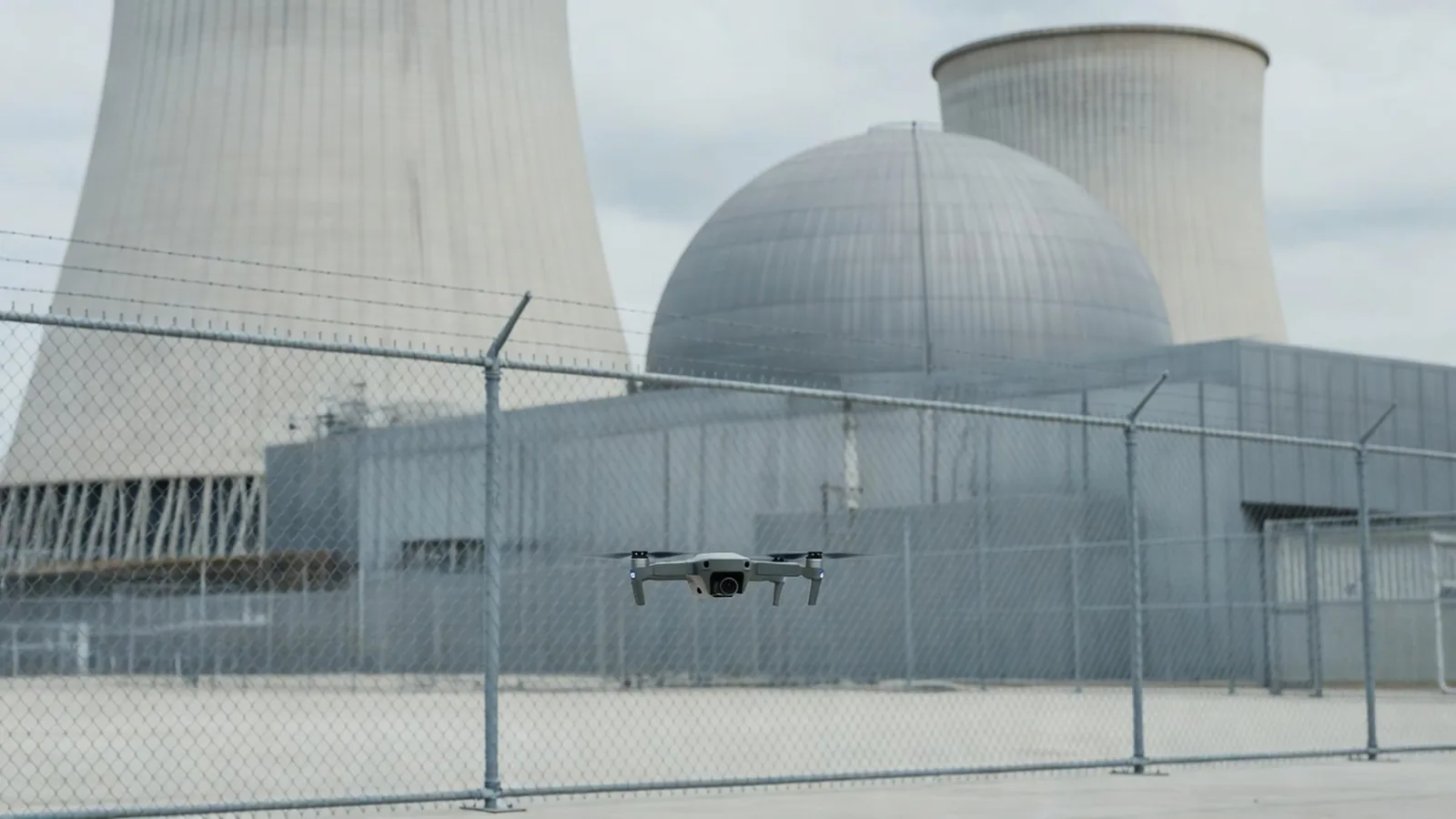 A small quadcopter drone hovering in the foreground with a nuclear power plant's cooling towers behind a security fence — legally observable, legally untouchable. Illustration: NNN.
