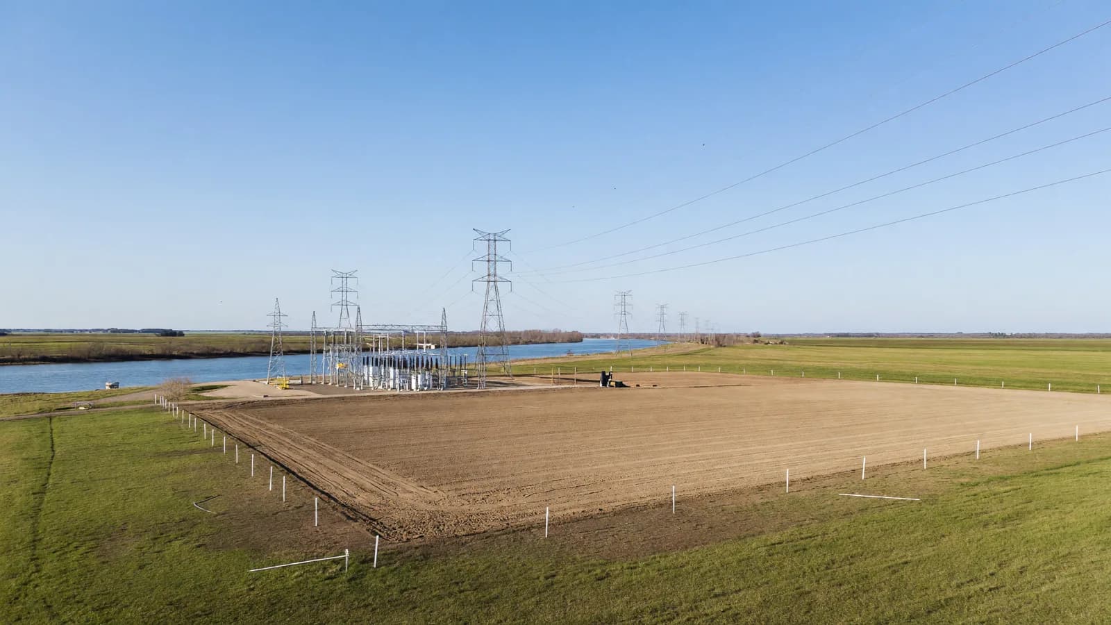 A cleared former coal-plant site on flat Indiana farmland beside a river, representing the retired Breed Generating Station where AEP, Amazon and X-energy are studying a 960 MW Xe-100 SMR plant. Illustration: NNN