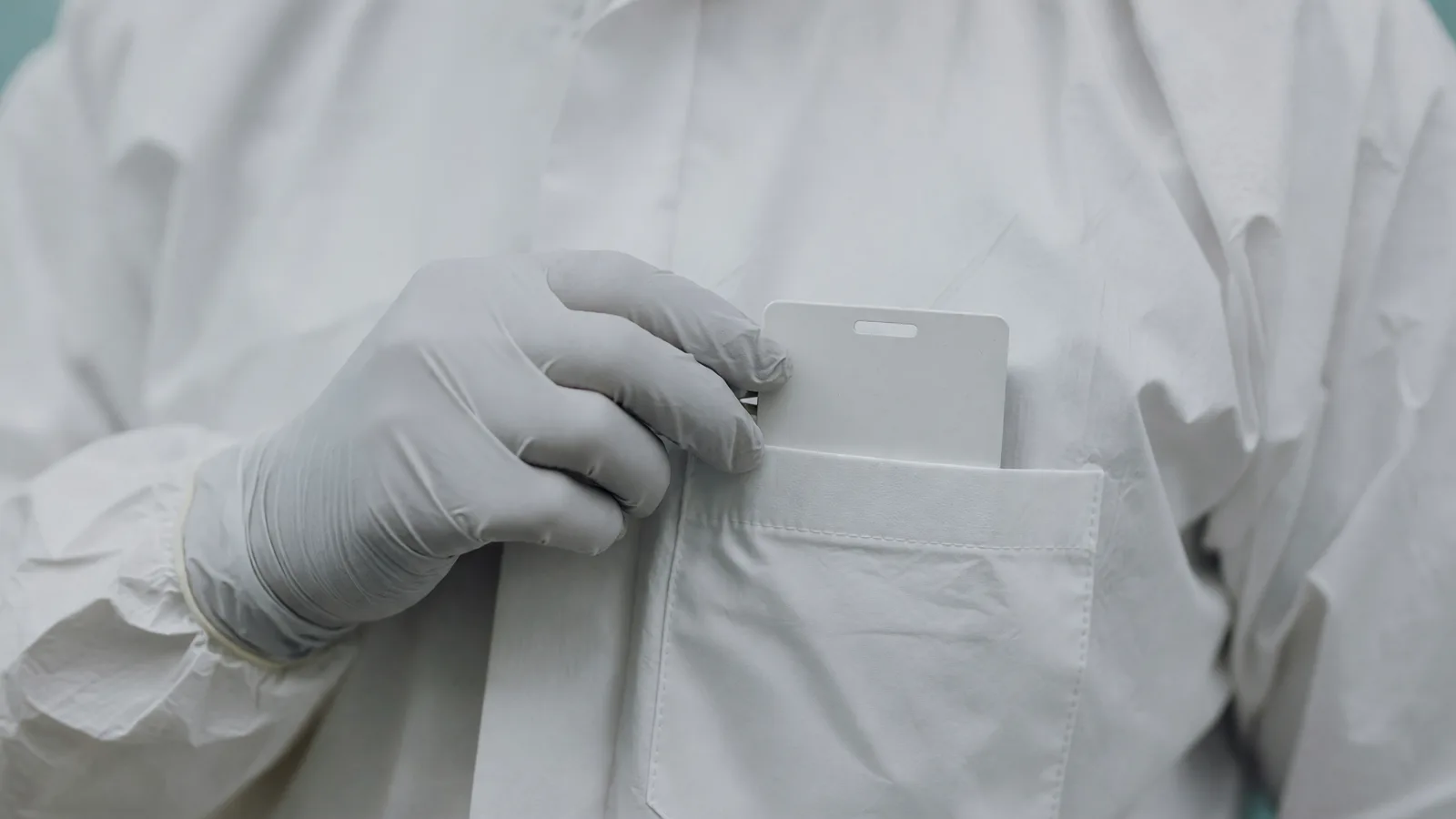 A gloved hand slips a dosimeter badge into a coverall chest pocket — the instrument at the centre of the ALARA rewrite. Illustration: NNN