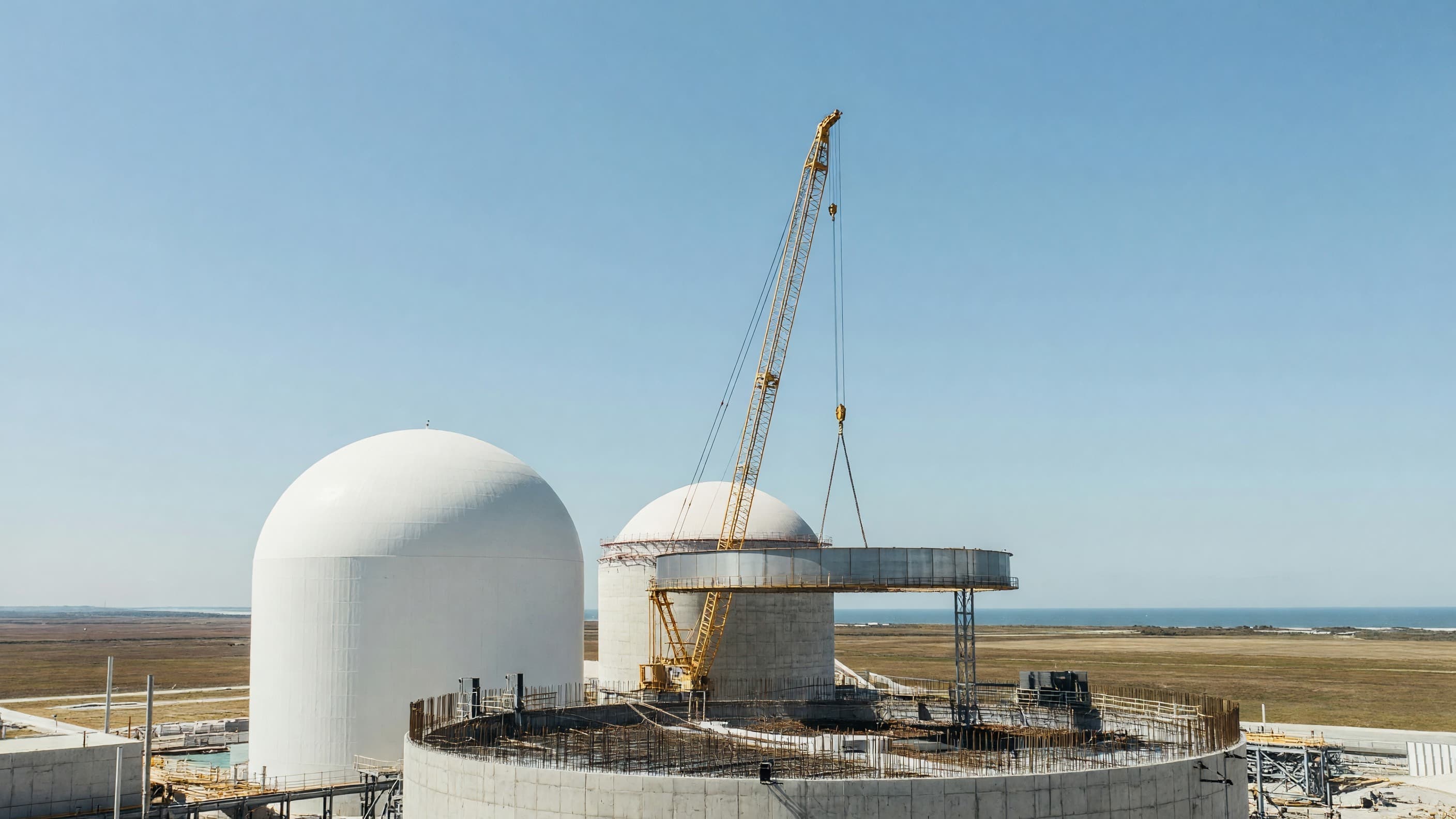 A pair of pressurized-water reactor containment buildings under construction at a US coastal nuclear site, one white containment dome complete and the second at ring-lift stage beneath a heavy-lift crane, illustrating the eight-reactor US build program South Korea is weighing as part of the tariff investment deal. Illustration: NNN