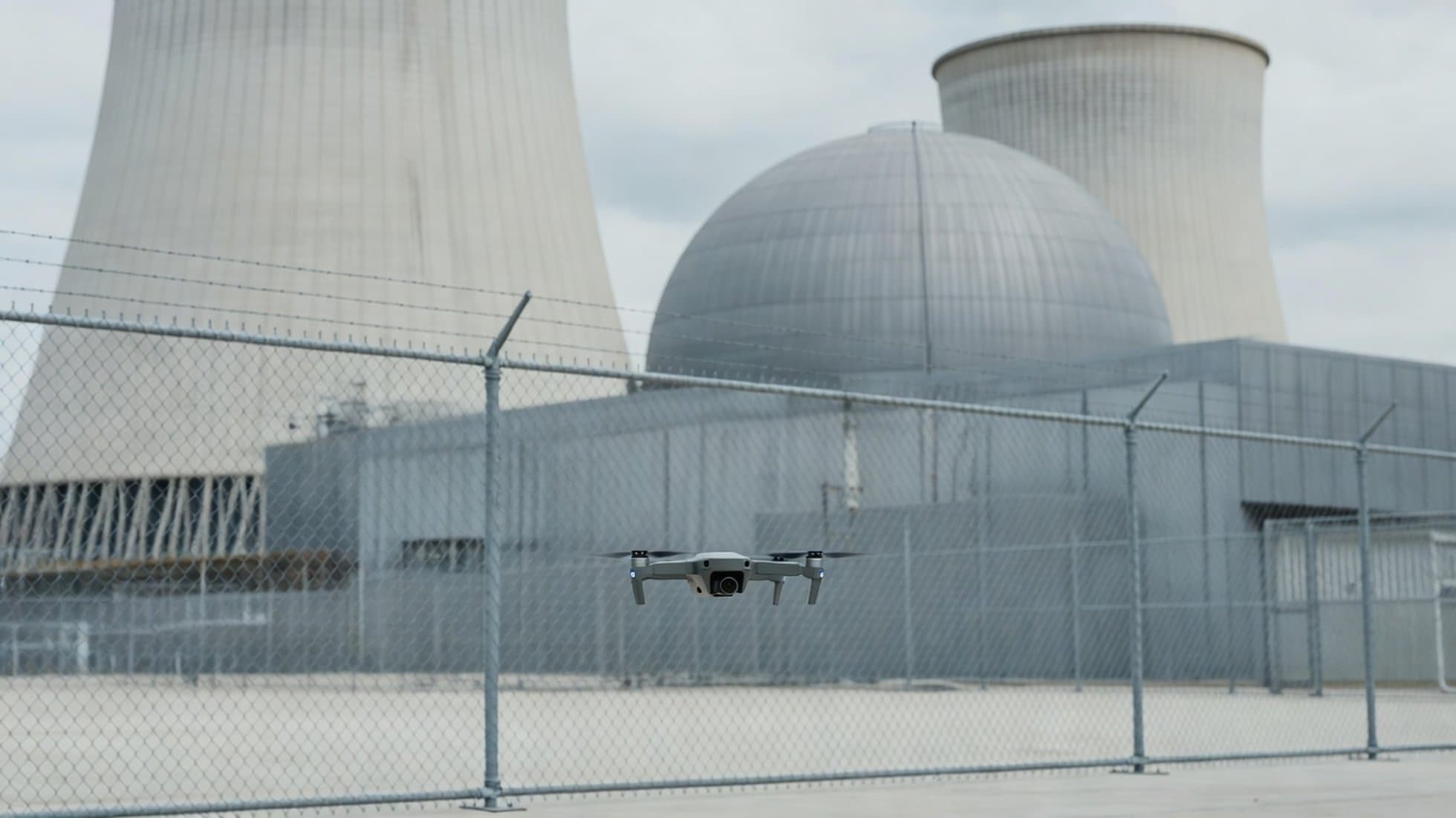 A small quadcopter drone hovering in the foreground with a nuclear power plant's cooling towers behind a security fence — legally observable, legally untouchable. Illustration: NNN.